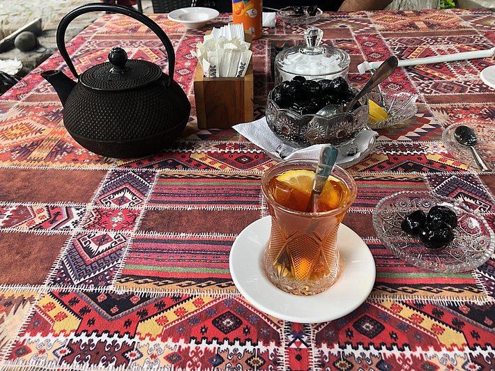 A beautifully arranged table features a traditional cast-iron teapot, a glass of tea with a slice of lemon on a white plate, and a dish of dark walnuts. The table is covered with a colorful patterned cloth, enhancing the inviting atmosphere.