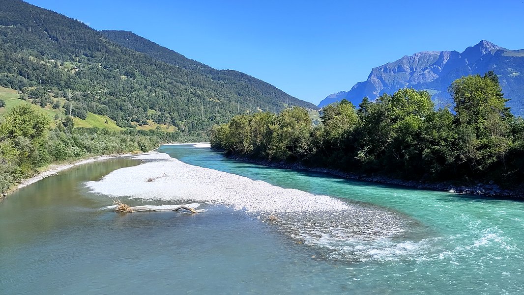 A river winds its way through a green landscape surrounded by trees and mountains. Small gravel islands are visible in the water, which shimmers in various shades of blue. The sky is clear and blue.