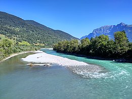Ein Fluss schlängelt sich durch eine grüne Landschaft, umgeben von Bäumen und Bergen. Im Wasser sind kleine Kiesinseln sichtbar, während das Wasser in verschiedenen Blautönen schimmert. Der Himmel ist klar und blau.