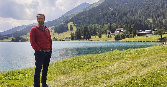A bearded man in a red jacket is standing in front of a lake in a mountainous landscape.