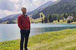 A bearded man in a red jacket is standing in front of a lake in a mountainous landscape.