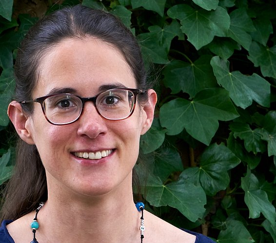 Woman with dark hair tied back in front of a wall covered with ivy wearing a dark blue blouse and a beaded necklace