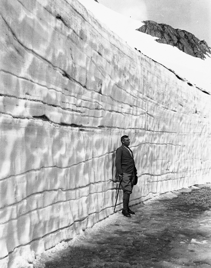 A man in a suit stands in front of a wall of snow that towers above him.