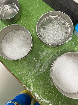 Verschiedene grobe Metallsiebe mit Schnee in unterschiedlichen Körnungen auf grünem Tisch