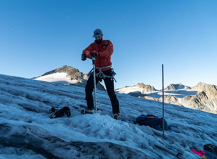 Person wearing a red jacket and black pants stands on a glacier using an ice auger to drill a hole in the ice. Mountain landscape in the background.