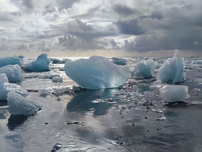 Eisberge schwimmen auf einer ruhigen Wasseroberfläche, umgeben von einer bewölkten Himmel. Die Eisschollen reflektieren das Licht und schaffen eine kühle, beeindruckende Landschaft.
