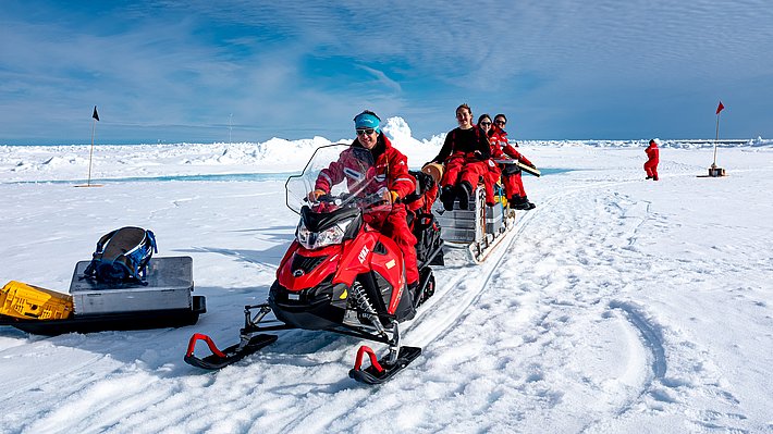 Eine Gruppe von Personen in roten Schneeanzügen sitzt auf einem Schlitten, der von einem roten Schneemobil gezogen wird. Im Hintergrund sind schneebedeckte Eisflächen und zwei Fahnen sichtbar.