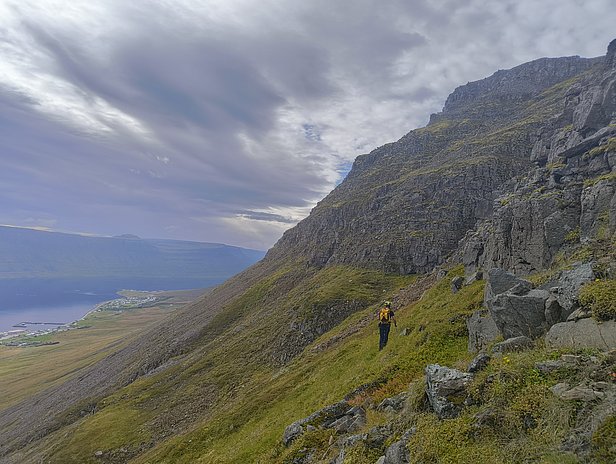 Ein Wanderer steht auf einem steilen, bewachsenen Hang mit Felsen. Im Hintergrund ist ein Tal und ein fjordartiger Gewässer sichtbar, umgeben von Bergen und Wolken. Die Szenerie vermittelt eine ruhige, natürliche Landschaft.