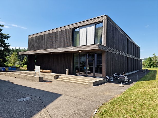 A modern wooden building with large windows, featuring a slight overhang and a set of steps leading to the entrance. Surrounding greenery and a clear blue sky can be seen in the background.