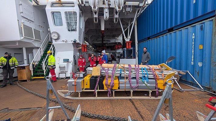Eine Gruppe von sieben Personen in roten und gelben Schutzanzügen steht auf einem Schiff. Im Vordergrund liegt eine große, gelbe Plattform mit mehreren Rohren und Seilen. Im Hintergrund sind Container und ein Steuerstand zu sehen.