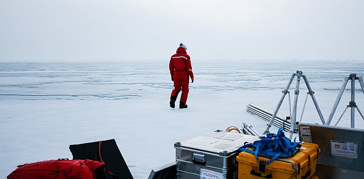 Person in rotem Overall geht auf zugefrorenen See, im Vordergrund Ausrüstungskisten und Stative auf Eisfläche.