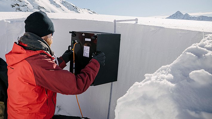 Un chercheur en vêtements d'hiver installe un appareil dans un trou de neige. La scène se déroule dans un environnement alpin, avec des montagnes enneigées en arrière-plan. L'action semble axée sur la collecte de données, soulignant l'importance de la recherche sur la neige.