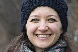 Eine junge Frau mit langen, braunen Haaren und einem freundlichen Lächeln steht vor einem neutralen Hintergrund. Sie trägt ein dunkelblaues Oberteil.