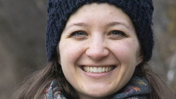 Eine junge Frau mit langen, braunen Haaren und einem freundlichen Lächeln steht vor einem neutralen Hintergrund. Sie trägt ein dunkelblaues Oberteil.