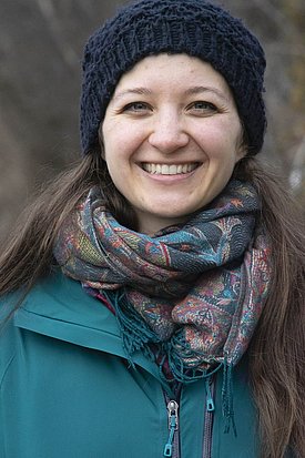 Eine junge Frau mit langen, braunen Haaren und einem freundlichen Lächeln steht vor einem neutralen Hintergrund. Sie trägt ein dunkelblaues Oberteil.