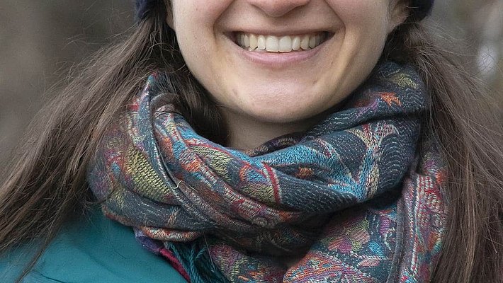 A woman with long dark hair smiles warmly at the camera. She is wearing a dark shirt and has a friendly demeanor, set against a plain, light-colored background.