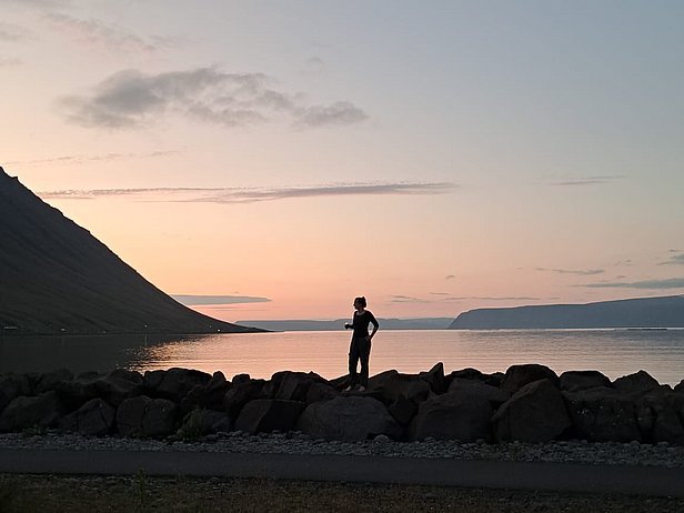 Une silhouette se tient sur une berge, face à un paysage de montagnes et d'eau douce. Le ciel est teinté de couleurs douces, indiquant le crépuscule. L'atmosphère est calme et sereine, évoquant un moment de contemplation.