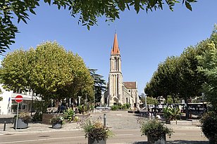 Platz im Genfer Vorort Lancy mit Bäumen, bepflanzten Behältern und einer Kirche mit rotem Turmspitzdach im Hintergrund