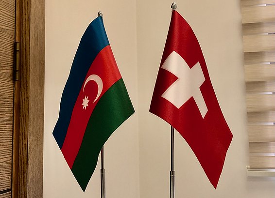 Two small flags are displayed on a table: the flag of Azerbaijan, featuring red, blue, and green stripes with a crescent and star, and the flag of Switzerland, which is red with a white cross. 