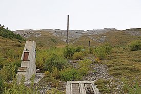 Eine verlassene Landschaft mit einer Betonstruktur im Vordergrund, umgeben von üppiger Vegetation. Im Hintergrund erhebt sich ein Berg mit sanften Hängen, und ein Stahlmast steht aufrecht in der Mitte der Szene, rechts davon ein kleinerer Mast. Der Himmel ist bewölkt und die Atmosphäre wirkt ruhig.