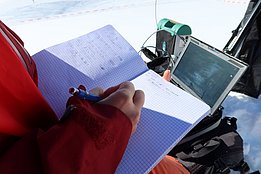 Anja Mödl in roter Skijacke schreibt in kariertes Notizbuch, im Hintergrund Laptop und Ausrüstung auf Schnee