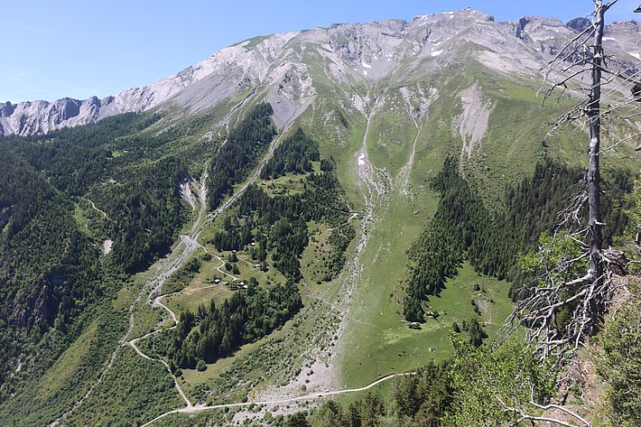 Ein Blick auf eine malerische Gebirgslandschaft mit grünen Wiesen und Wäldern. Steile Hänge und Felsen bilden die Kulisse, während sich kurvenreiche Wege durch die Landschaft schlängeln. Der Himmel ist klar und blau, was die natürliche Schönheit der Umgebung verstärkt.