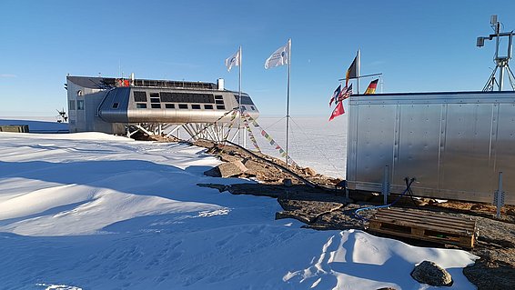 Forschungsstation auf schneebedecktem Berggipfel mit mehreren Flaggen unter klarem blauem Himmel