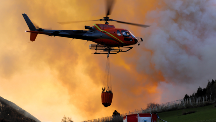 Elicottero con serbatoio rosso antincendio vola davanti a un fumo arancione sopra un campo verde con una tenda dei vigili del fuoco.