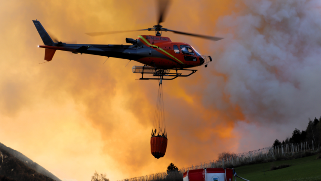 A helicopter carrying a red water tank flies over a green field where there is a tent belonging to the fire brigade, with orange smoke visible in the background.
