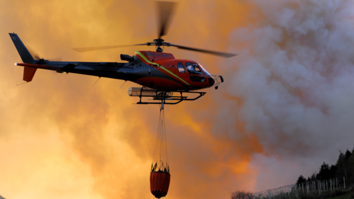 Hubschrauber mit rotem Löschbehälter fliegt vor orangefarbenem Rauch über grünem Feld mit Feuerwehrzelt.