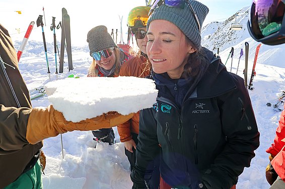 Eine Gruppe von vier Personen steht im Schnee. Eine Hand hält ein großes Stück kompakten Schnees und ein Frau schaut es an. Die anderen schauen interessiert zu. Im Hintergrund sind Skier und Berge sichtbar.