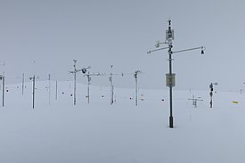 Messstation mit verschiedenen Geräten steht in einer schneebedeckten Landschaft unter grauem Himmel