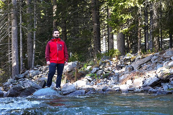 Ein Mann steht in einem roten Jacket am Ufer eines Flusses, umgeben von Bäumen und Felsen. Das Wasser fließt ruhig im Vordergrund, während die Landschaft im Hintergrund grün und natürlich wirkt.