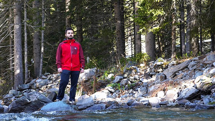 Ein Mann steht in einem roten Jacket am Ufer eines Flusses, umgeben von Bäumen und Felsen. Das Wasser fließt ruhig im Vordergrund, während die Landschaft im Hintergrund grün und natürlich wirkt.