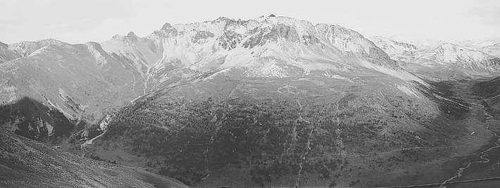 Schwarz-weiss Foto: Schneebedeckte Berggipfel mit steilen Hängen und einem bewaldeten Tal im Vordergrund.