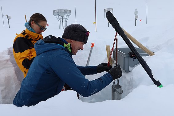 Zwei Personen in Winterkleidung bestimmen Schneedichte in einer Schneegrube mit Messgeräten.
