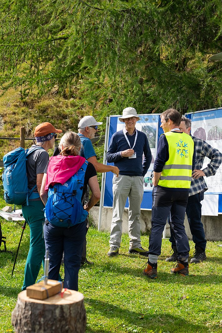 Mehrere Personen stehen auf einer Wiese, umgeben von Bäumen. Eine Person trägt eine gelbe Warnweste, andere haben Rucksäcke. Im Hintergrund sind Informationsplakate.