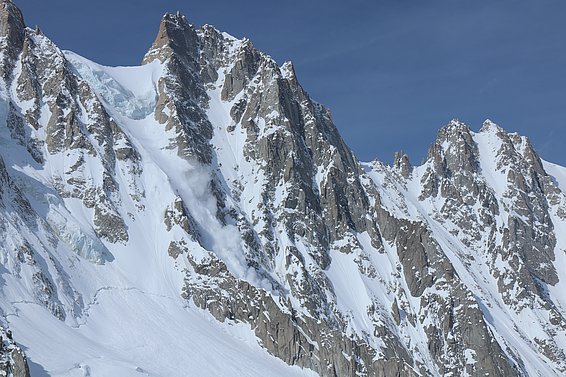 A dramatic mountain landscape features steep, jagged peaks covered in snow. An avalanche can be seen cascading down one side, sending a plume of snow and ice into the air. The blue sky provides a stark contrast to the rugged, white terrain.