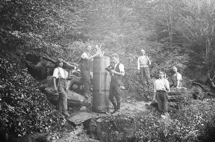 A group of men are standing in the forest around a bin with branches sticking out of it.