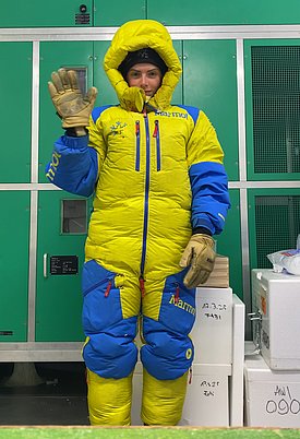 Mira Lange in gelb-blauem, dick gepolstertem Expeditionsanzug winkt mit Handschuhen vor grünen Schränken