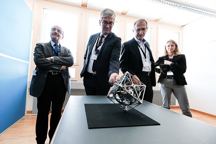 Ein Würfel aus Metall und viel Elektronik steht auf einer seiner Ecken und wird von vier Personen betrachtet