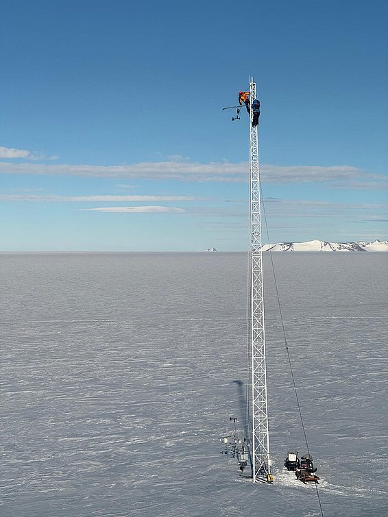 Zwei Personen in Schutzkleidung arbeiten an einer hohen Metallmessstation in einer weiten, schneebedeckten Landschaft mit Bergen im Hintergrund.