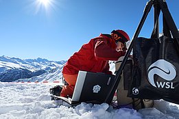 Anja Mödl in rotem Skianzug kniet im Schnee und schließt ein Spektrometer an einen Laptop an, im Hintergrund verschneite Berge und blauer Himmel.