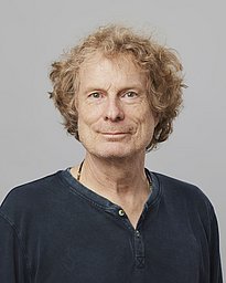 A middle-aged man with curly, light brown hair and a slight smile is wearing a dark-colored, long-sleeve shirt. He has an approachable expression against a neutral background.