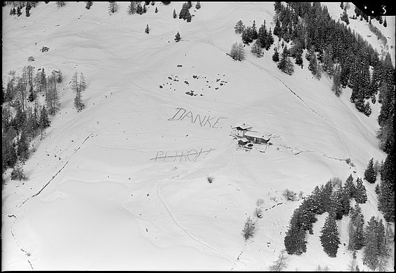 Vue aérienne en noir et blanc d'un paysage montagneux enneigé avec des arbres, une petite cabane et les mots 'DANKE' ainsi qu'un mot barré écrits dans la neige