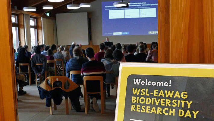 Une salle de conférence remplie de participants assis, regardant un présentateur au fond. Un panneau en premier plan indique "Bienvenue ! Journée de recherche sur la biodiversité WSL-EAWAG". L'ambiance est professionnelle, axée sur des discussions scientifiques.