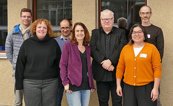 A group of seven individuals stands together, smiling. They display a mix of formal and casual attire. The background features a beige wall with windows. The group appears diverse in gender and age, suggesting a collaborative or professional setting.