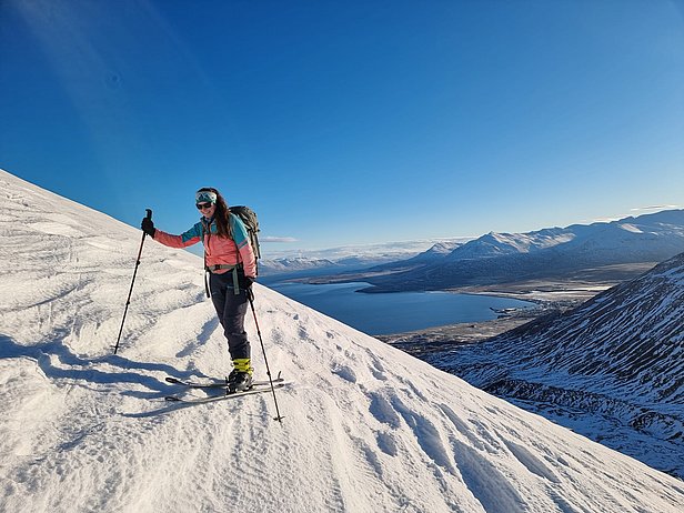 Eine Person in Skibekleidung steht auf einem schneebedeckten Hang und hält einen Skistock. Im Hintergrund erstreckt sich ein See und die umliegenden Berge unter klarem, blauem Himmel.
