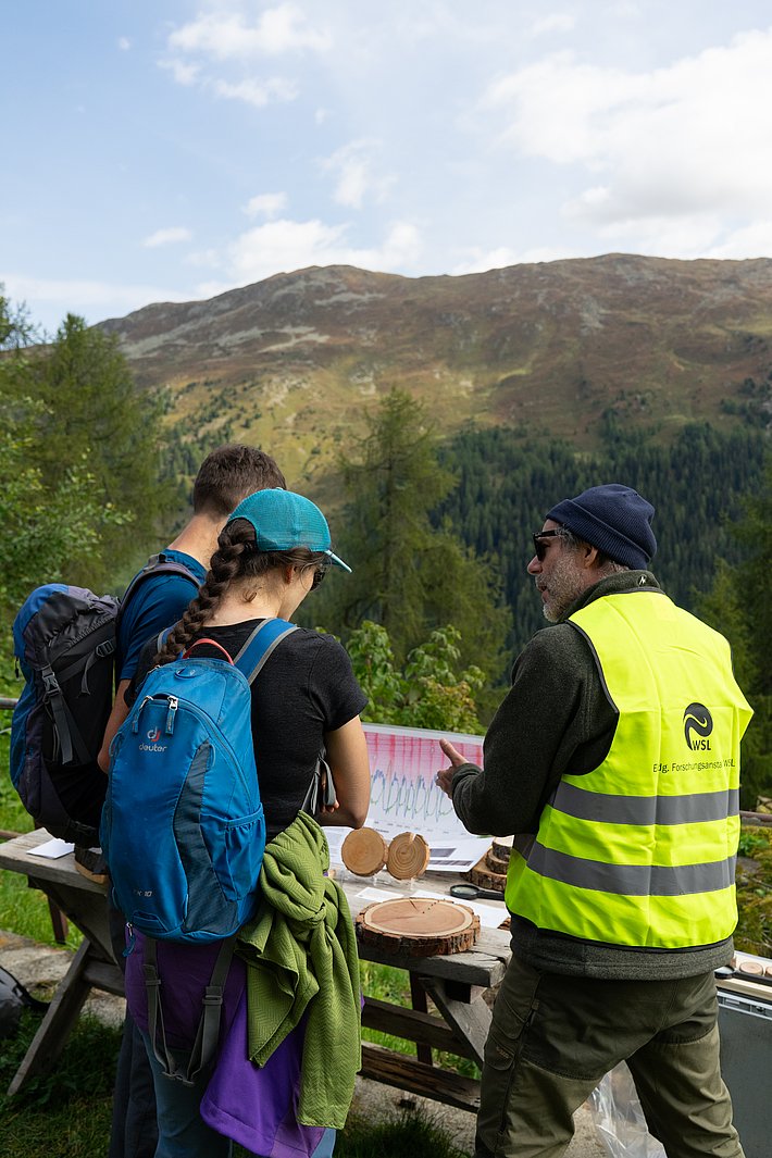 Drei Personen stehen an einem Tisch im Freien, der mit Holzscheiben und Materialien zum Lernen über Natur ausgestattet ist. Im Hintergrund sind Berge und Bäume sichtbar.