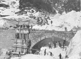 A group clears a railway bridge buried by an avalanche. Some of the group are standing under the bridge, some are on the bridge, and some are on a wooden scaffold on the bridge.